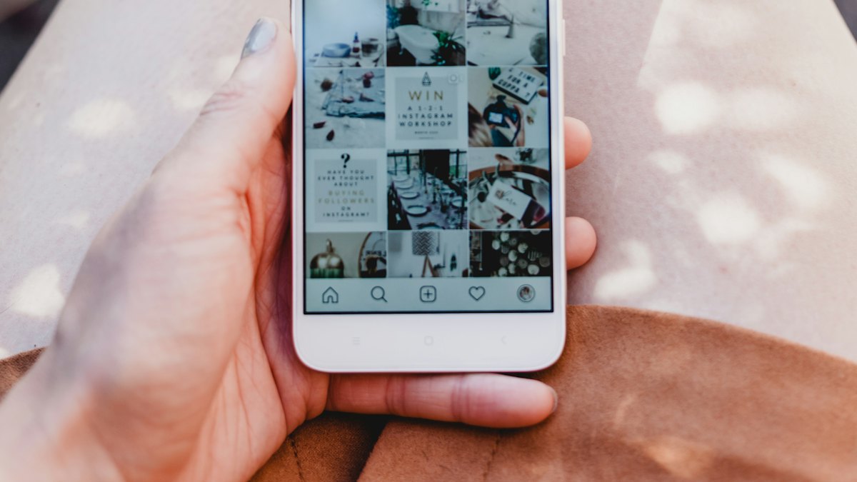 A hand holding a phone showing an Instagram profile grid