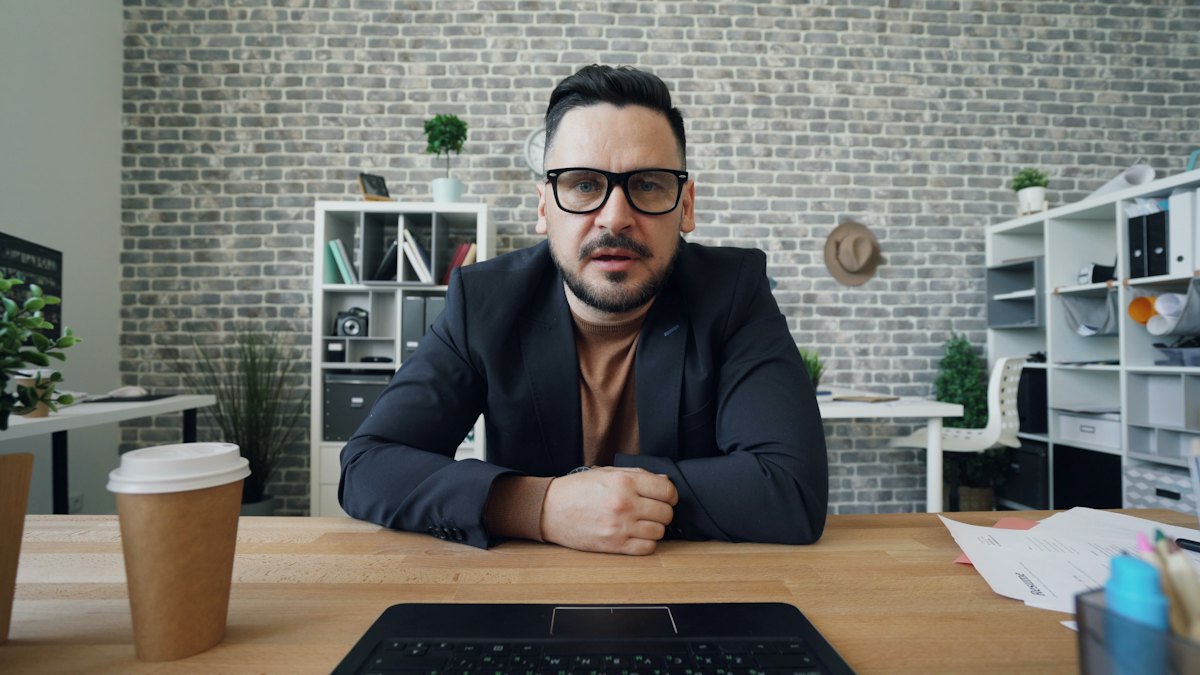 A man in a jacket talking to a laptop camera in an office